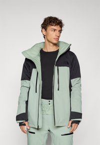 Jachetă impermeabilă, de culoare verde mentă și neagră, cu glugă, dotată cu buzunare cu fermoar și blocuri de culoare contrastante. Textură netedă și design modern.
