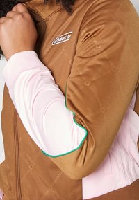 Person wearing a brown Adidas jacket with subtle embossed pattern, pink sleeves, and green piping, with arm bent and hand near chin.