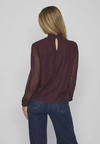 Blusa bordeaux con maniche lunghe e trasparenti caratterizzata da un motivo a zigzag, colletto alto e dettaglio a chiave sul retro. Abbinata a jeans in denim scuro.
