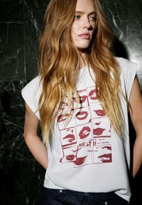 Wit mouwloos shirt met een rood lippenstiftprintontwerp, met een raster van lippen en de tekst "BORN IN LONDON" en "WEST 11" eronder.