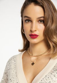 Maglione beige a V impreziosito con paillettes argentate, abbinato a collane in oro a strati. Il modello ha capelli ondulati e labbra rosa audaci.