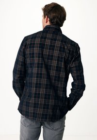 Camicia a scacchi blu navy con dettagli marroni e bianchi, maniche lunghe, colletto abbottonato e orlo leggermente curvo. Indossata con jeans grigi.