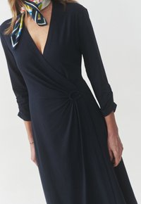Navy wrap dress in soft fabric, featuring 3/4 sleeves, a twist detail at the waist with a circular accent, and a v-neckline.