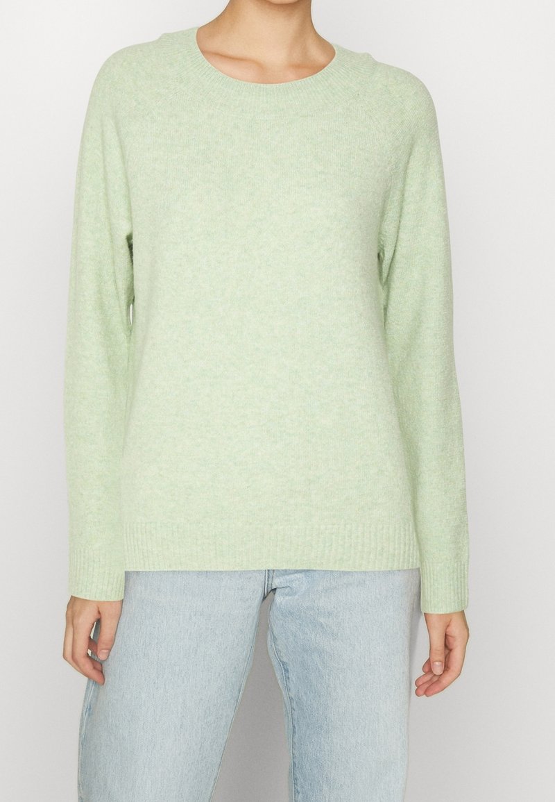 Maglione verde chiaro con scollatura rotonda, maniche raglan lunghe e fondo a coste. Tessuto morbido; indossato sopra jeans azzurri chiari.