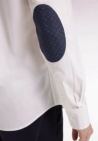 Chemise blanche à manches longues avec une épaule texturée marine comportant un motif bleu subtil. Poignets boutonnés, coupe ajustée, tissu lisse.