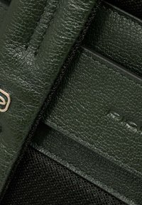 Cuero texturizado de color verde oscuro con un acabado suave, con un detalle de logo en tono dorado y detalles en tela negra. Paneles verticales mostrados.