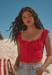 Top corto rojo con tejido texturizado y mangas acampanadas. Presenta un detalle de lazo en la parte delantera, combinado con pantalones cortos de denim de cintura alta.