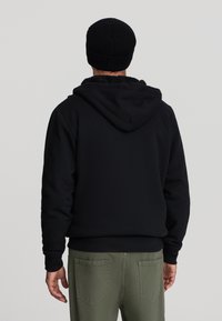 Felpa nera con cappuccio, foderata in morbido tessuto, polsini a coste e tasca a marsupio, abbinata a pantaloni verde oliva e a un berretto di maglia nero.