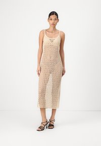 Knit dress in light beige with an open weave design, featuring thin straps and a mid-calf length. Paired with black strappy heeled sandals.