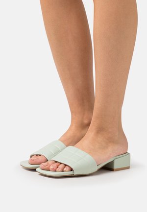 Piedi che indossano sandali aperti con tacco basso a blocco in verde chiaro trapuntato su uno sfondo bianco.