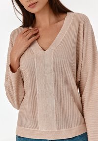 Pull en tricot beige clair avec un col en V et une texture côtelée. Dispose de longues manches amples et d'une coupe droite.