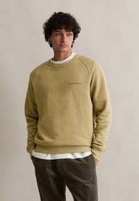 Lichtgroene sweatshirt met geribbelde mouwen, ronde halslijn en een tekstenlogo op de borst. Draag het over een witte shirt en gecombineerd met donker gestreepte broek.