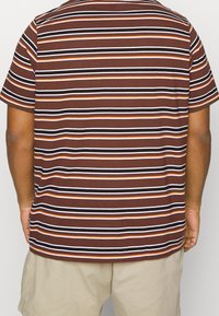 Persona con una camisa de rayas horizontales en marrón, naranja, blanco y negro, de manga corta, y pantalones cortos beige, vista desde la espalda.