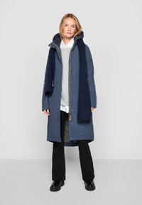 Marine parka met een gestructureerde afwerking, rits en drukknoopjes, een capuchon en een contrasterende gele rits. Draagt over een grijs sweater en zwarte broeken.