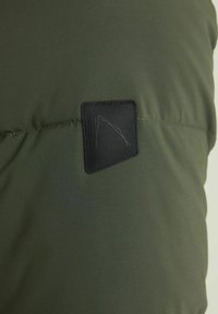 Primo piano di una giacca imbottita verde con tessuto strutturato, una toppa nera rettangolare con un logo e dettagli di cuciture visibili.