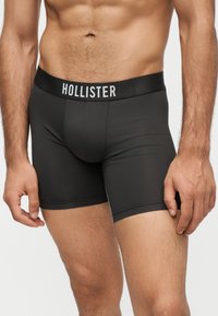 Άνδρας με μαύρα μποξεράκια με το λογότυπο "Hollister" στη μέση, στέκεται με τα χέρια χαλαρά στο πλάι.