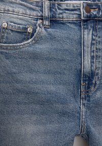 Section avant d'un jean en denim bleu montrant la ceinture, la boucle de ceinture, le bouton métallique et la petite poche à monnaie avec rivets.