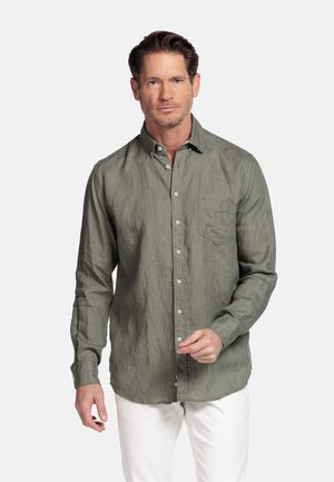 Homme portant une chemise à manches longues boutonnée vert olive et un pantalon blanc, debout devant un fond clair uni.