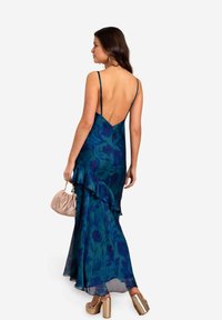 Abito lungo senza maniche, con scollo a V profondo sulla schiena, di colore blu scuro con motivi floreali, orlo a volant, realizzato in tessuto leggero, abbinato a una borsa color beige.