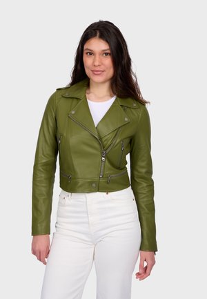 Vrouw draagt een olijfgroene leren jas met ritssluitingdetails en witte broek, staand tegen een effen lichte achtergrond.