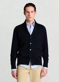Cardigan côtelé bleu marine avec col châle, boutonné sur le devant, et manches longues, porté sur une chemise à rayures bleu clair. Texture douce.