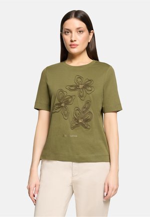 Kobieta ubrana w oliwkowo zielony krótki top z trzema haftowanymi kwiatami i napisem "Flower Garden", w połączeniu z jasnobeżowymi spodniami.