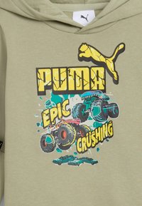 Felpa khaki con logo PUMA giallo screpolato, due camion mostruosi colorati che schiacciano auto color teal, e la frase "EPIC CRUSHING" in caratteri in grassetto.