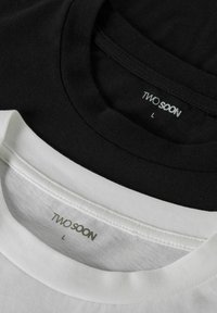 Due t-shirt, una nera e una bianca, impilate con scolli rotondi. Le etichette riportano "TWO SOON" e la taglia "L" in un font semplice.