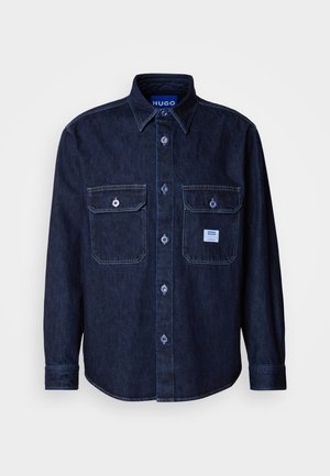 Mørk denim skjorte med knapper, lange ærmer, to brystlommer og blå mærketag. Har kontrastsyning og klassisk krave.