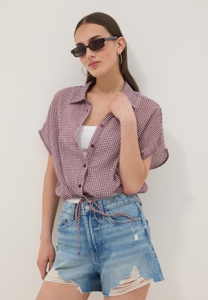 Giovane donna con occhiali da sole rettangolari neri, orecchini a cerchio, camicia rossa a quadri annodata, canottiera bianca e shorts in denim slavati.