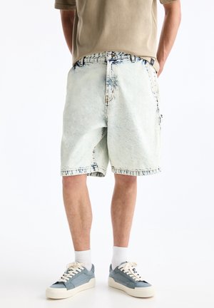 Homme portant un short en jean délavé clair, des chaussettes blanches, des baskets grises et une chemise beige, debout avec les mains croisées derrière le dos.