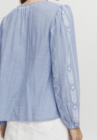 Vaaleansininen pusero, joka on tehty teksturoidusta kankaasta, jossa on valkoisia kirjailtuja kuvioita hihojen ja kaula-aukon ympärillä. Hihoissa on rypytetty hihansuu ranteessa.