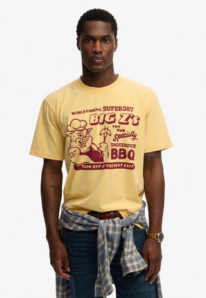 Mężczyzna w żółtej koszulce z bordowym motywem i napisem BBQ, kraciastą koszulą zawiązaną wokół talii, niebieskich dżinsach i zegarku na lewej ręce.