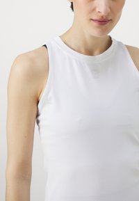 Balta sporta krekls bez piedurknēm ar gludu tekstūru, apaļu izgriezumu kakla vietā un mazu logotipa detaļu uz krūtīm. Iekšpusē ir melna siksniņa.