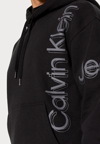 Sudadera negra con el logotipo plateado de Calvin Klein en vertical en el frontal y parte del logotipo en la manga, con un bolsillo tipo canguro y cordones.