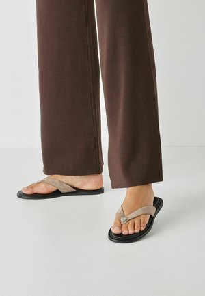 Brune flip-flops med en beige ruskind V-formet rem, der har en sølvfarvet hjerteaccent og en sort gummisål, kombineret med løse brune bukser.