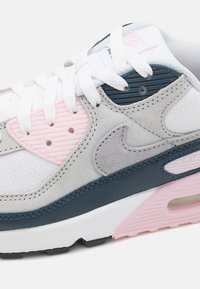 Zapatilla blanca con gamuza gris y detalles en azul. Presenta un revestimiento de plástico rosa, paneles texturizados y cordones blancos. Amortiguación de aire visible.