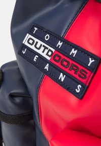 Punainen ja tummansininen vedenpitävä laukku, jossa on suorakulmainen brodeerattu logo, jossa lukee "TOMMY OUTDOORS JEANS" valkoisella ja mustalla tummansinisellä taustalla.