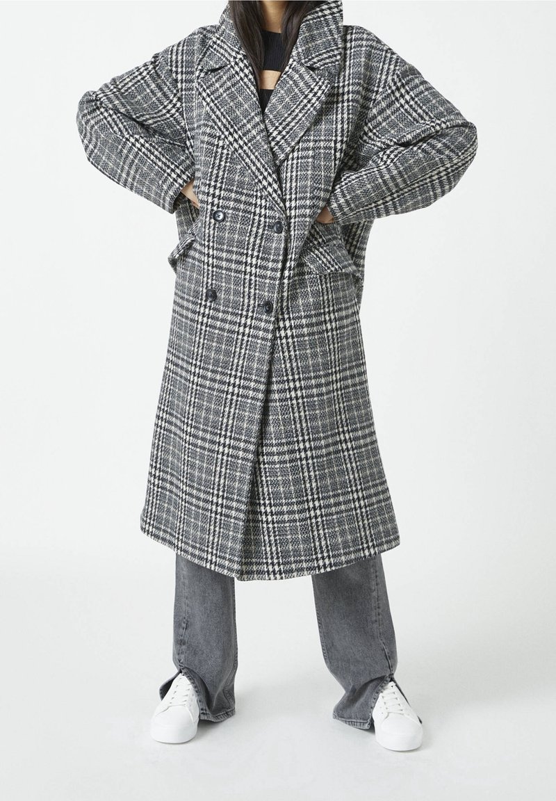 Harmaa-valkoinen ruudullinen takki, jossa kaksirivinen nappilista, suuret kaulukset ja sivutaskut, kerroksittain harmaiden farkkujen ja valkoisten tennarien päällä.