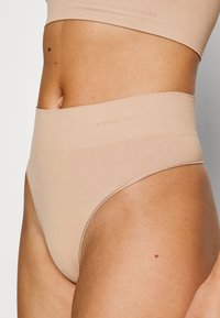 Beige seamless thong underwear with a high waistband and smooth texture, featuring an embossed logo on the waistband.