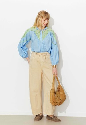 Femme portant un chemisier bleu clair avec de la dentelle verte, un pantalon beige à jambes larges, des mocassins marron, tenant un sac à main en daim marron.