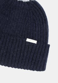 Beanie lavorato a maglia blu navy con una texture a coste, dotato di un piccolo etichetta bianca con la scritta "AX" cucita vicino al polsino e un morbido pom-pom sulla parte superiore.