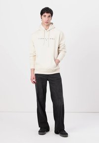 Crème hoodie met het "TOMMY JEANS" logo, een voorzak en een koord. Gecombineerd met donkergrijze wijde jeans en zwarte schoenen. Minimalistisch ontwerp.