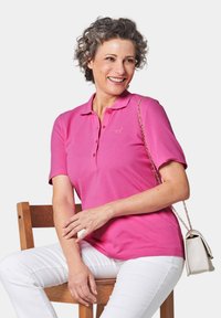 Roze polo shirt met korte mouwen, een drieknoopsluiting en geborduurd logo. Gecombineerd met witte broeken en een kleine crèmekleurige crossbody tas.