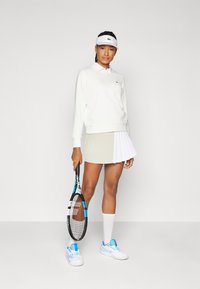 Tenis obleka z belim puloverjem, s srajco s ovratnikom, plisirano bež in belo krilo, belimi nogavicami in teniškimi čevlji z modrimi akcenti, ki drži lopar.