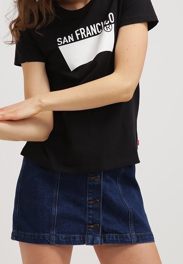 Ung person klädd i en svart t-shirt med texten "San Francisco" och en blå knäppt denimkjol, med armarna korsade framför sig mot en neutral bakgrund.