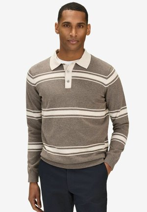 Mand iført en brun langærmet polotrøje med hvide vandrette striber og mørke bukser, stående med den ene hånd i lommen.