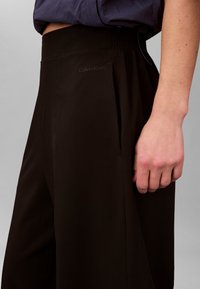 Άτομο που φοράει μαύρο παντελόνι Calvin Klein με ψηλή μέση, ορατή ζώνη και πλαϊνή τσέπη, το χέρι ακουμπά κοντά στον γοφό, σε απλό φόντο.