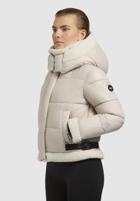 Beige puffjakiga, millel on vale karusnaha krae ja mansetid, vööd ning küljetaskud. Tekstuuriga pind ja tikitud disain.