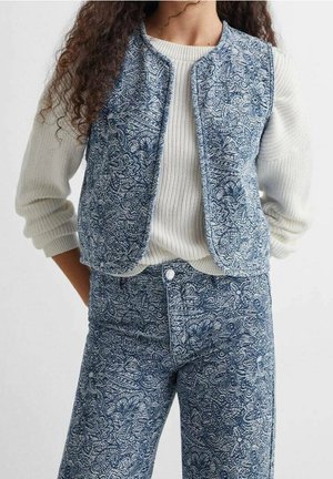 Femme portant un pull côtelé blanc avec manches retroussées, assorti à un gilet bleu à motif floral et un pantalon taille haute coordonné.